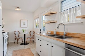 Une cuisine moderne avec des meubles blancs, un plan de travail en bois, un évier encastré et une fenêtre avec des rideaux. On y trouve deux chaises en osier disposées autour d’une petite table sur laquelle est posé un vase de fleurs.