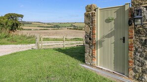A rustic stone building with a green door, a small window with a light fixture, and a view of a rural landscape with fields and a tree. - Garstons Barn (Newport)