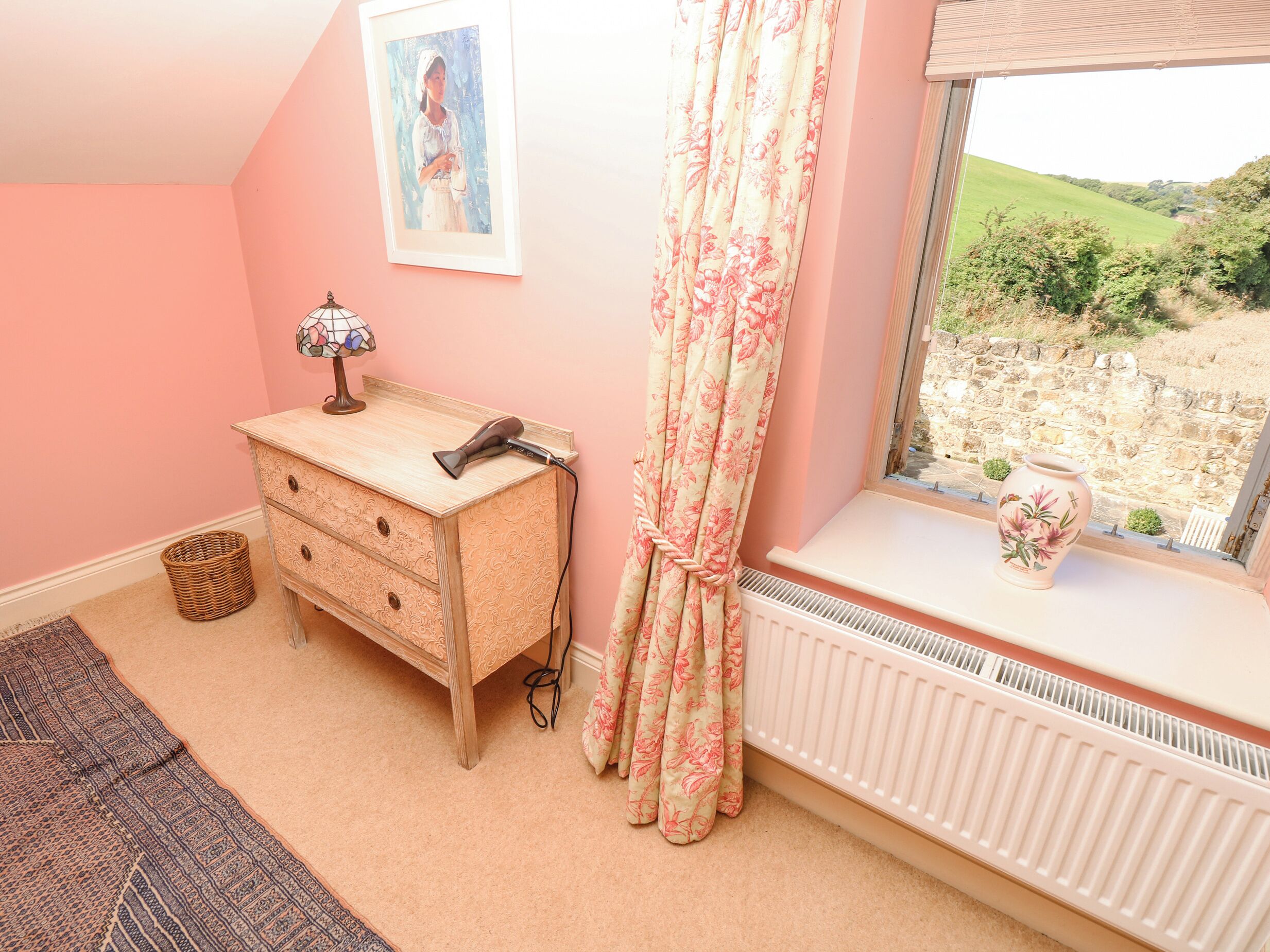 A bedroom with a dresser, a radiator, a window with a view of greenery, and a lamp.