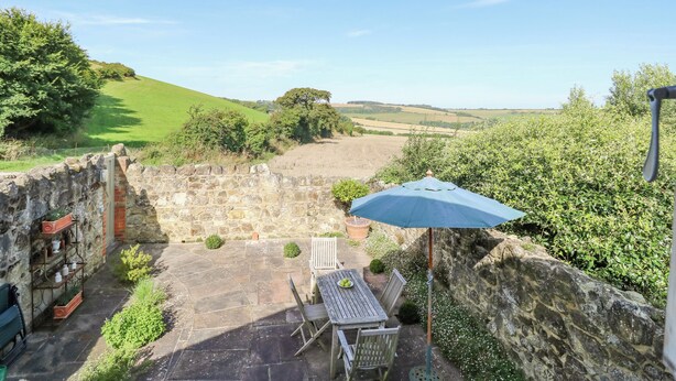A stone wall, a table with chairs, and a blue umbrella in a garden with a view of rolling hills. - Garstons Barn (Newport)