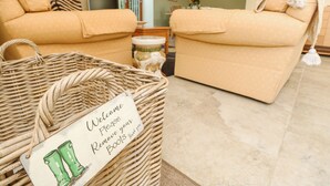A cozy living room with two beige sofas, a wicker basket with a 'Welcome Please Remove your Boots Thank you' sign, and a dining area in the background. - Garstons Barn (Newport)
