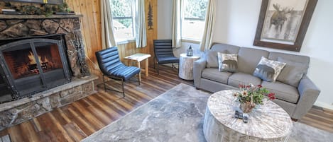 A cozy living room with a fireplace, wooden flooring, a sofa, a coffee table, and a view of the outdoors.