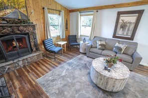 A cozy living room with a fireplace, wooden flooring, a sofa, a coffee table, and a view of the outdoors. - Lazy R Cottages: 7a 2 Bedroom Cabin (Estes Park)