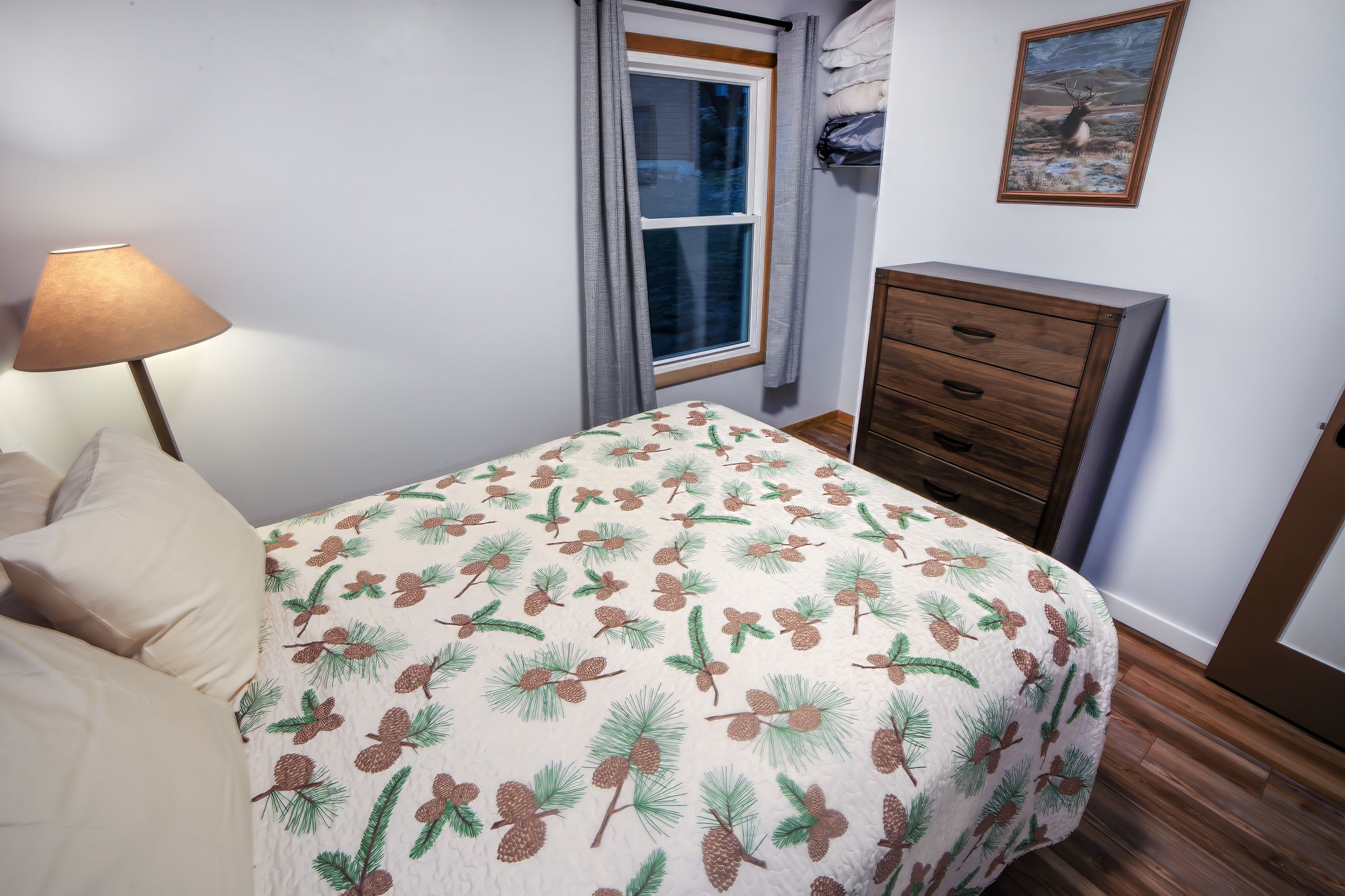 Ein Schlafzimmer mit einem Bett, einem Nachttisch, einer Lampe, einem Fenster mit Vorhängen, einer Kommode und einem Bild an der Wand.