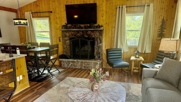 A cozy living room with a stone fireplace, wooden walls, a round coffee table, and a comfortable seating area. - Lazy R Cottages: 7a 2 Bedroom Cabin (Estes Park)