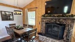 A dining area with a wooden table and chairs, a fireplace, and a mounted TV. - Lazy R Cottages: 7a 2 Bedroom Cabin (Estes Park)