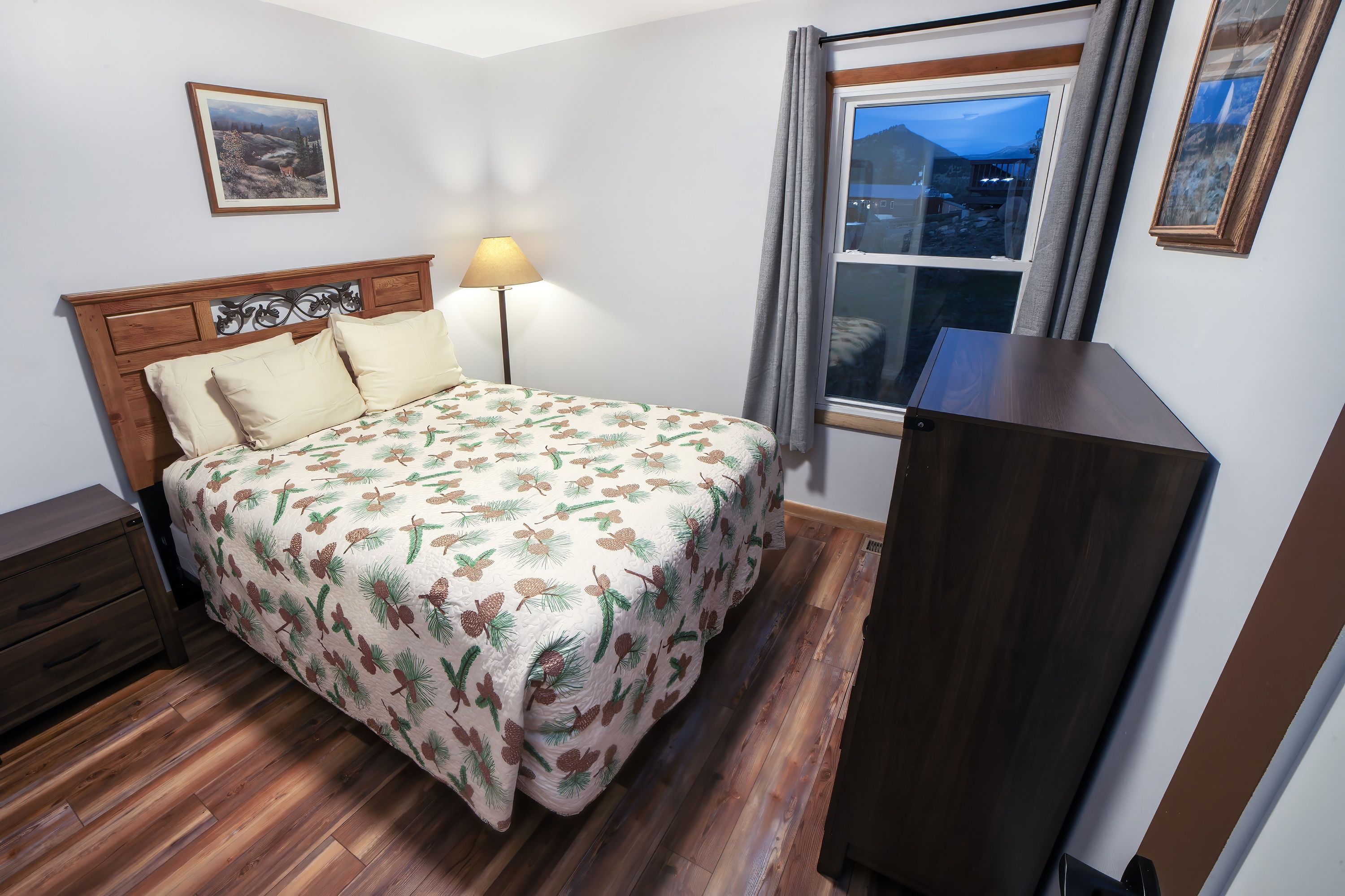A bedroom with a bed, a nightstand, a lamp, a window with curtains, and a framed picture on the wall.