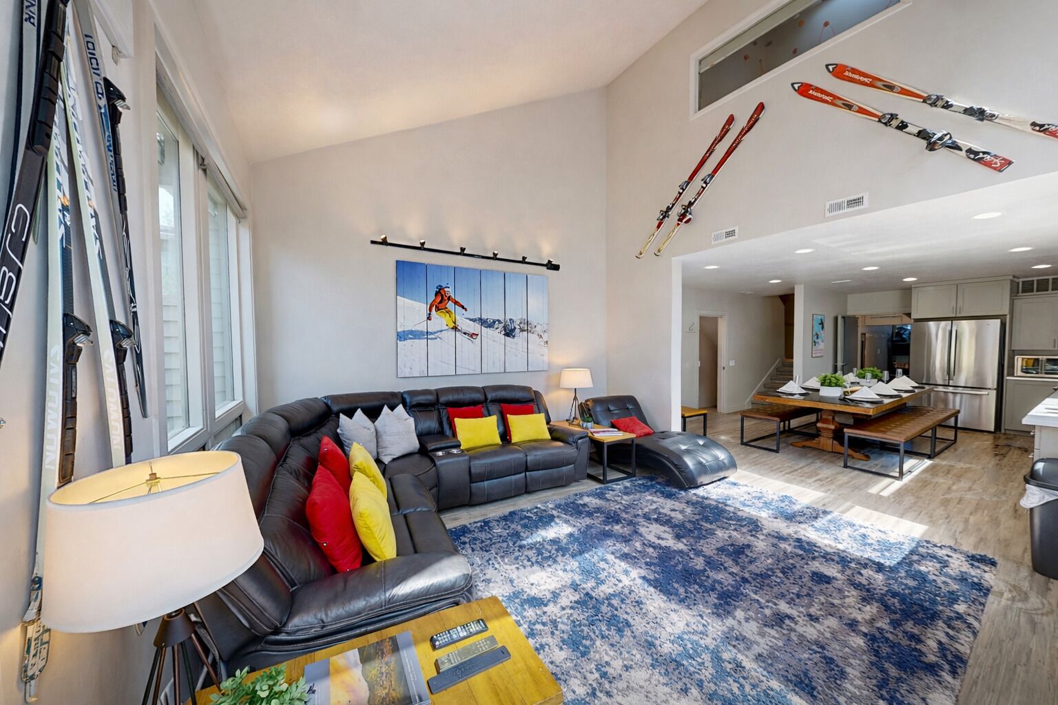 A modern living room with a black leather sofa, colorful cushions, a blue patterned rug, and a dining area with a wooden table and chairs.