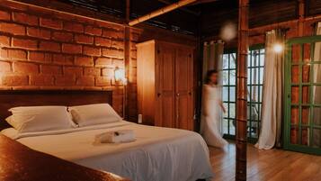 Quarto com parede de tijolos, vigas de madeira no teto, cama com lençóis brancos e porta verde.