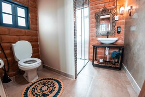 A bathroom with a white toilet, a sink with a mirror, and a patterned rug on the floor. - Bangalô Ametista (Alto Paraíso de Goiás)