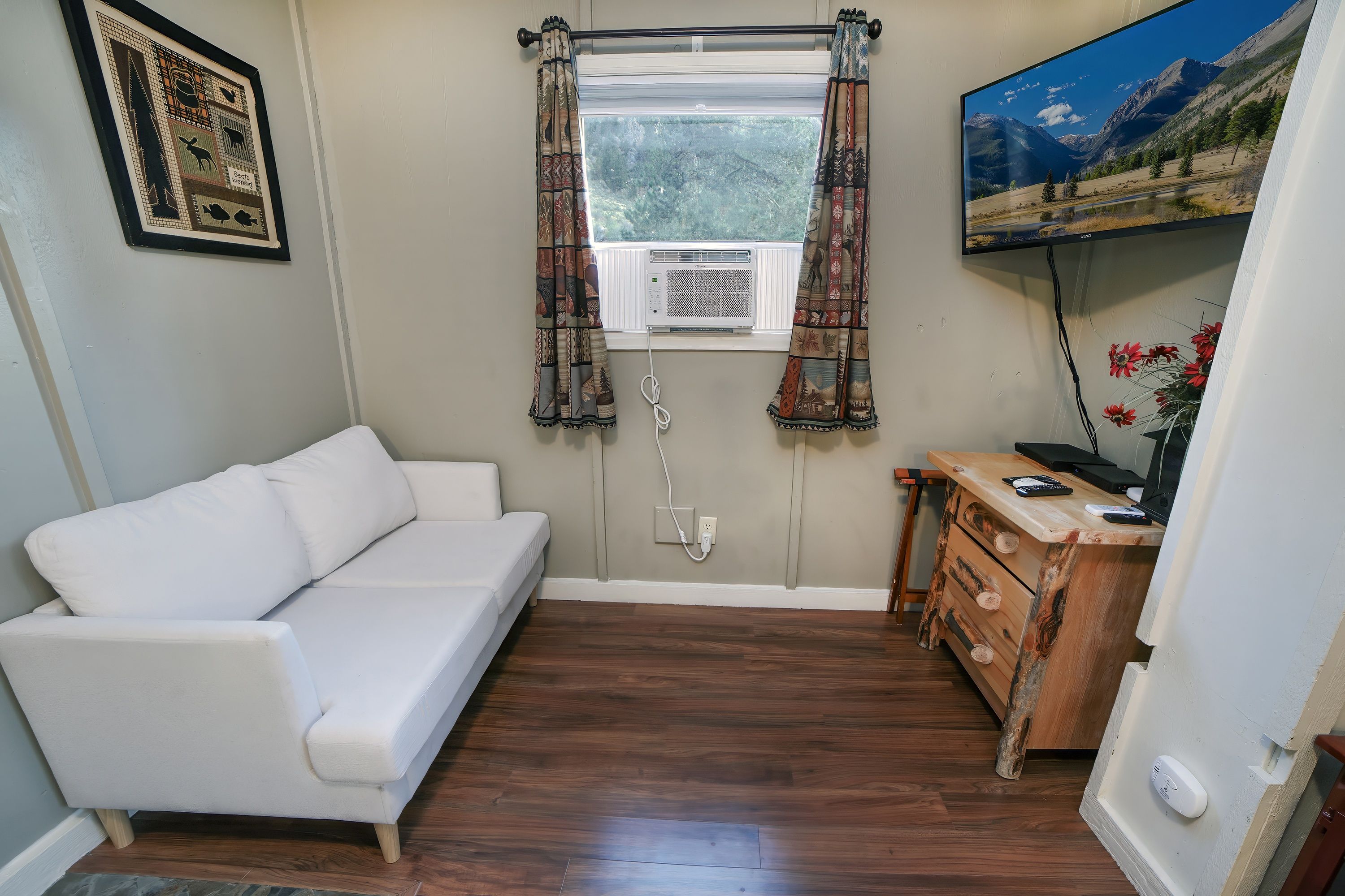 Una habitación con un sofá blanco, un aparador de madera, un televisor montado y una ventana con cortinas estampadas.