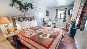 A cozy bedroom with a large bed, a wooden headboard, a chair, a small table, and a TV. - Fawn Valley Inn: Cabana 1 Bedroom Apts (Estes Park)