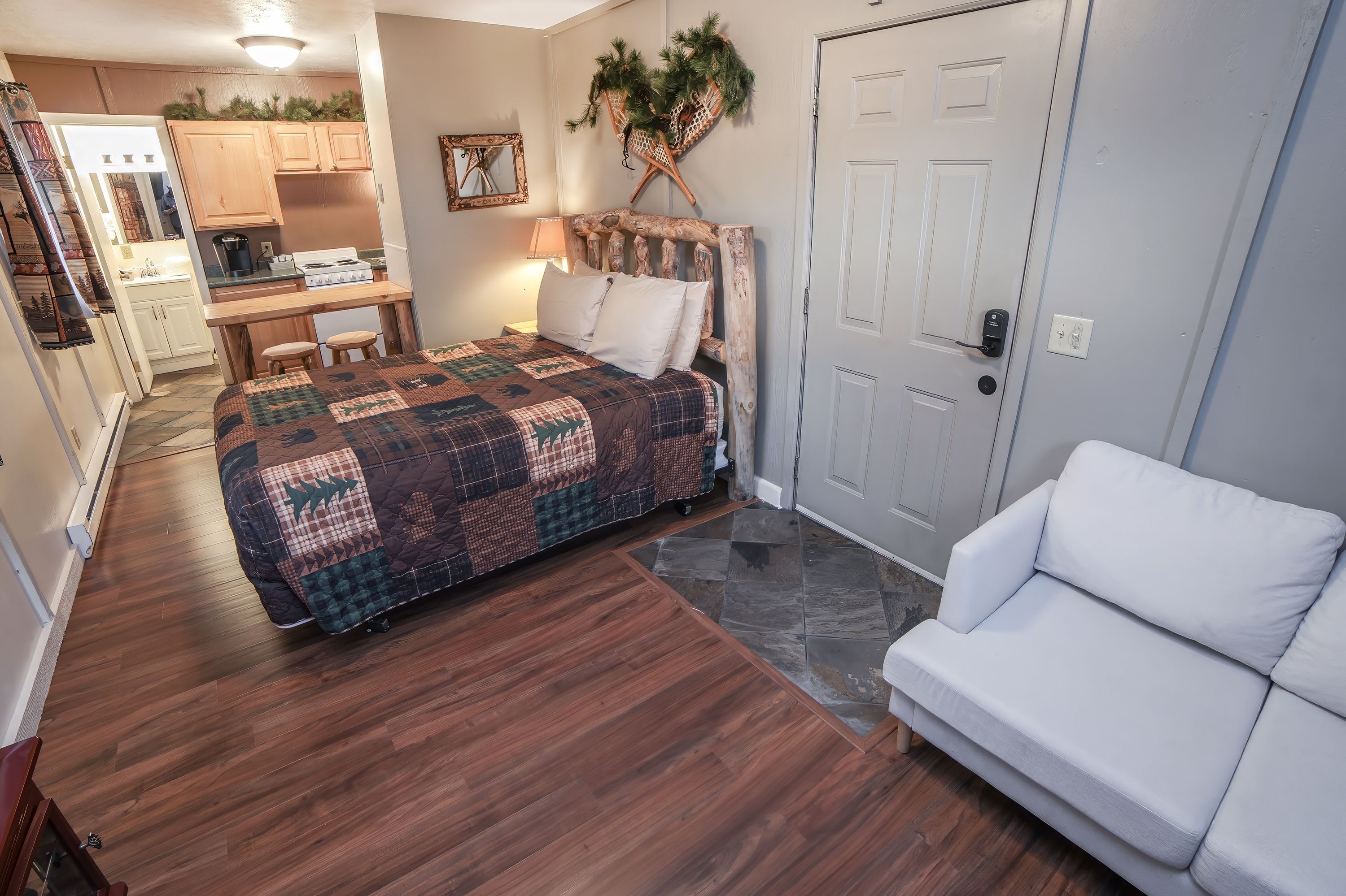 A bedroom with a bed, a white sofa, a wooden desk with a chair, a kitchen area, and a door with a lock.