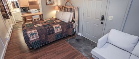 A bedroom with a bed, a white sofa, a wooden desk with a chair, a kitchen area, and a door with a lock.