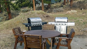An outdoor dining area with a round table and wooden chairs, surrounded by two barbecue grills. - Fawn Valley Inn: Cabana 1 Bedroom Apts (Estes Park)