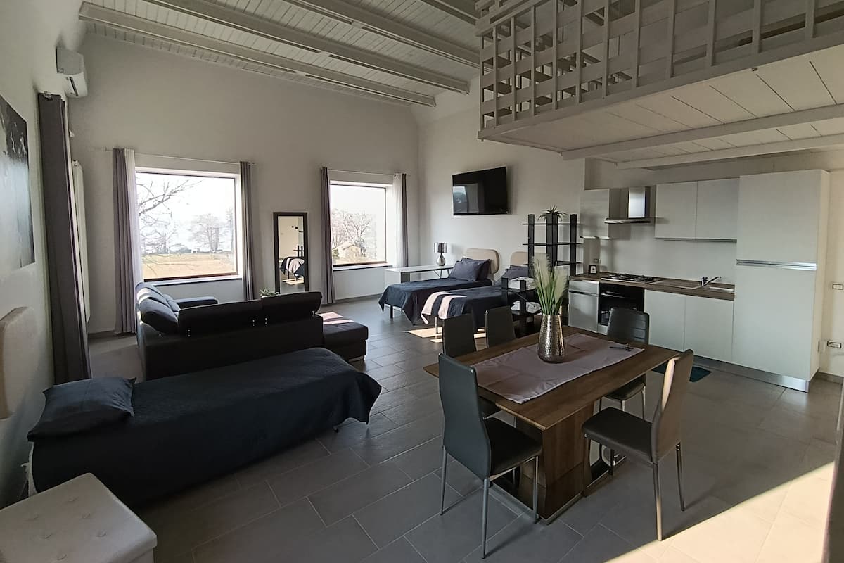 A modern living room with a sofa, a dining table with chairs, a kitchen area, and a large window.
