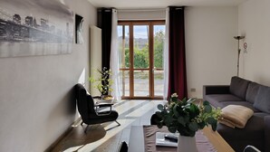 Un salón moderno con un sofá, un sillón, una mesa con un jarrón, un radiador y un cuadro de un paisaje urbano.