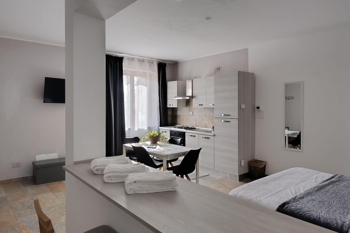 A modern hotel room with a bed, a dining table with chairs, a kitchen area, and a large mirror.
