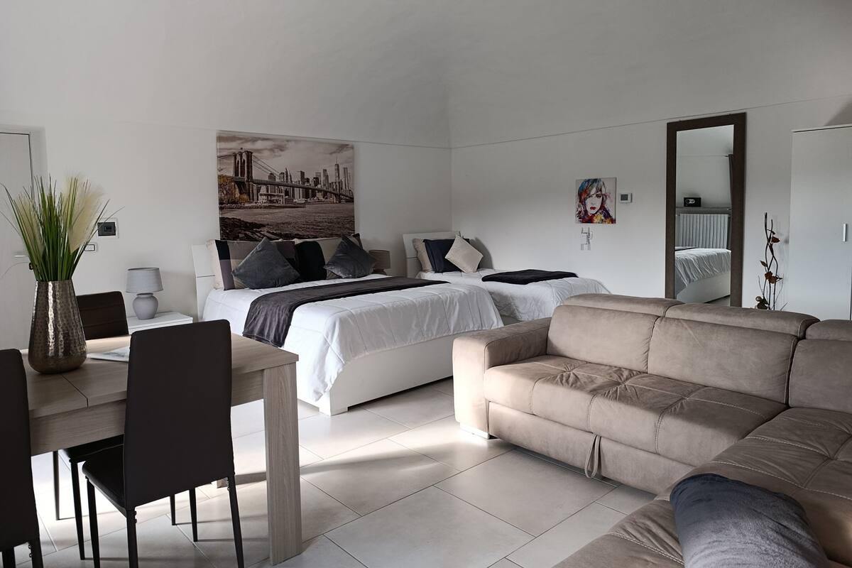 A modern bedroom with a bed, a sofa, a dining table with chairs, and a large mirror.