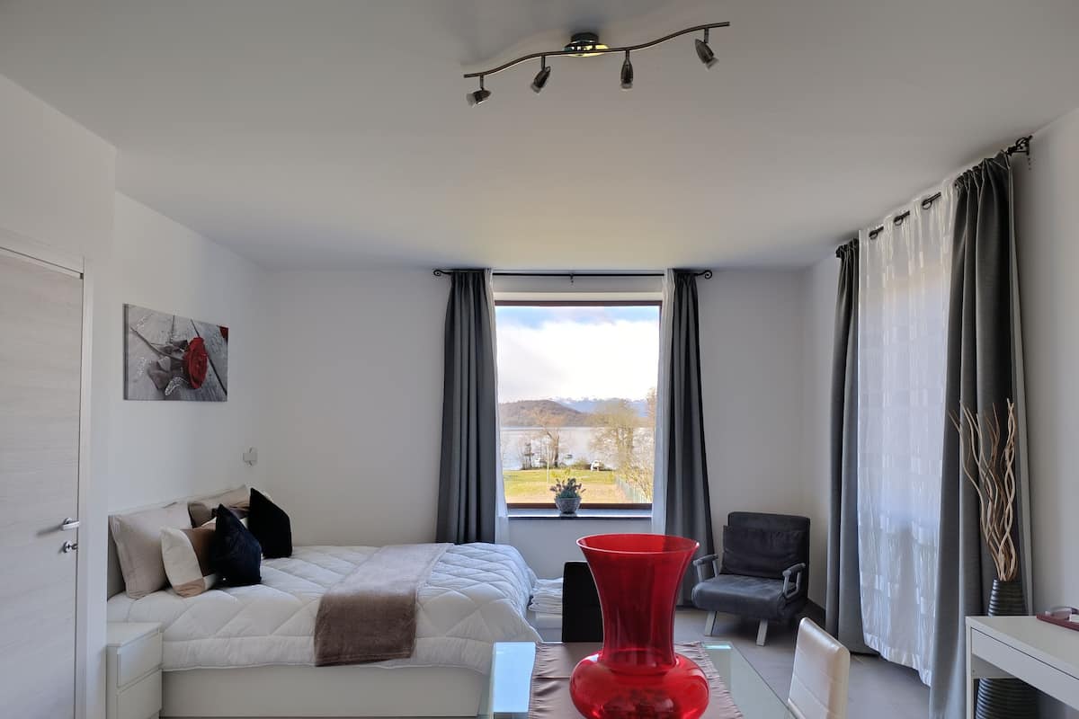 A bedroom with a bed, a red vase, a chair, and a view of a lake.