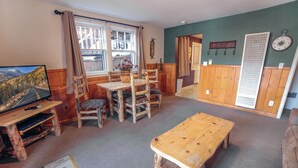 Een gezellige kamer met een houten tafel en stoelen, een televisie en een groot raam met gordijnen.
