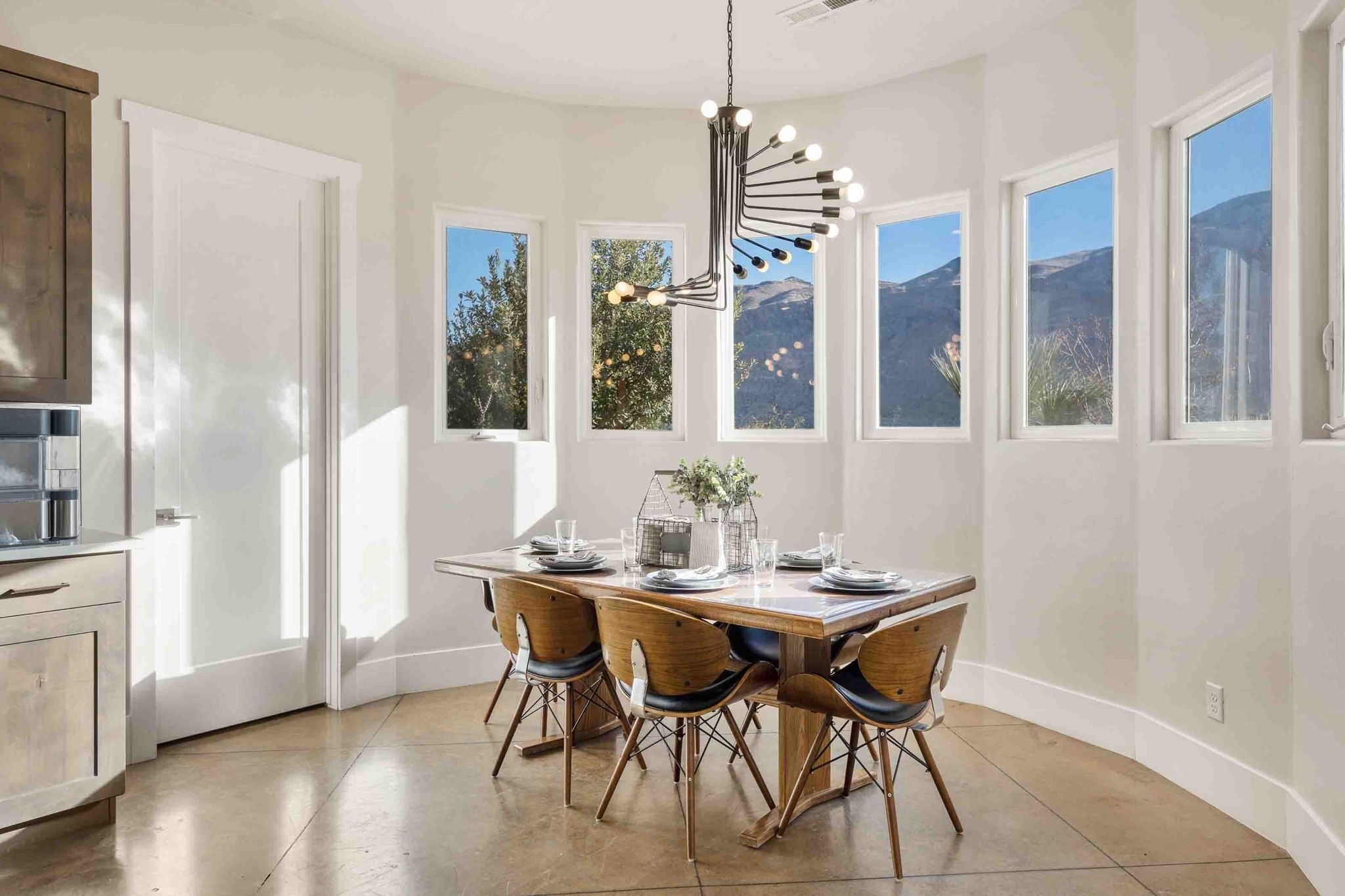 A dining area with a wooden table and chairs, large windows with a mountain view, and a modern chandelier.