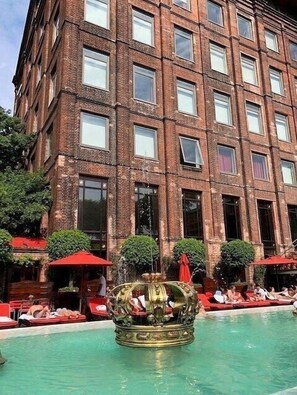 Un gran edificio de ladrillo con numerosas ventanas y un área de piscina con sombrillas rojas y sillones de descanso.