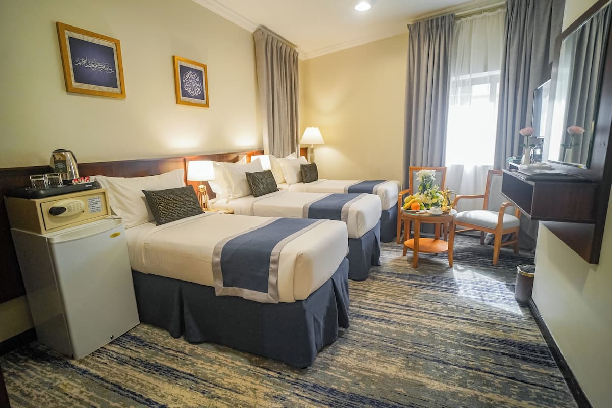 Habitación de hotel con dos camas, un escritorio con lámpara, un refrigerador pequeño, una silla y una ventana con cortinas.