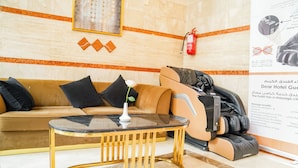 Un vestíbulo de hotel con un sofá, una silla de masaje, una mesa con superficie de vidrio y un extintor en la pared.
