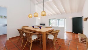 Un comedor con mesa de madera y sillas de mimbre, una cocina al fondo y vista al exterior por una ventana.