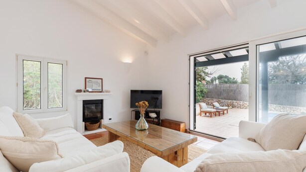 Amplia sala de estar con mobiliario blanco, chimenea, televisor y un gran ventanal que da a un patio exterior.
