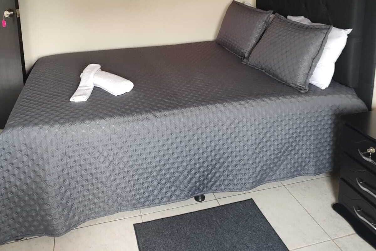 A neatly made bed with a grey quilt, a folded towel, and a black headboard.