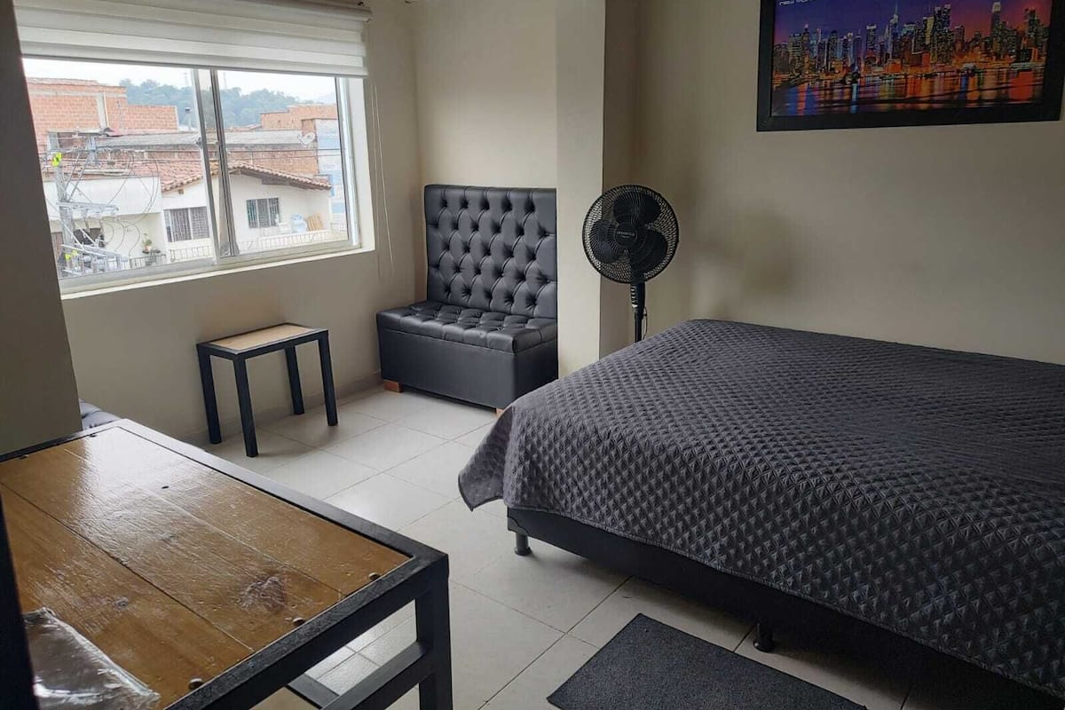 A bedroom with a bed, a chair, a table, a standing fan, and a window with a cityscape view.