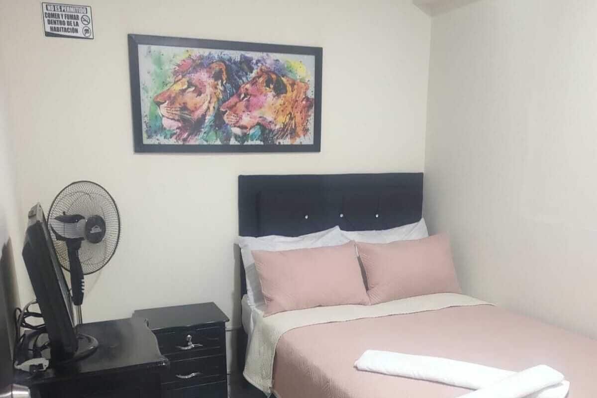 A bedroom with a bed, a desk with a chair, a standing fan, and a framed painting on the wall.