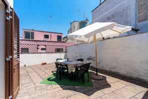 Eine Terrasse mit Tisch, Stühlen, einem weißen Sonnenschirm und einer grünen Matte.