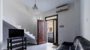 Un salon avec un canapé en cuir noir, un téléviseur posé sur un meuble noir, un escalier et une porte donnant sur l’extérieur.