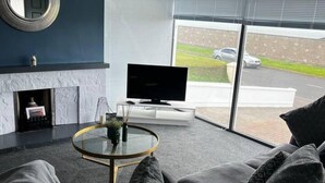 Una sala de estar moderna con chimenea, televisor de pantalla plana y un ventanal con vistas a un estacionamiento y un automóvil.