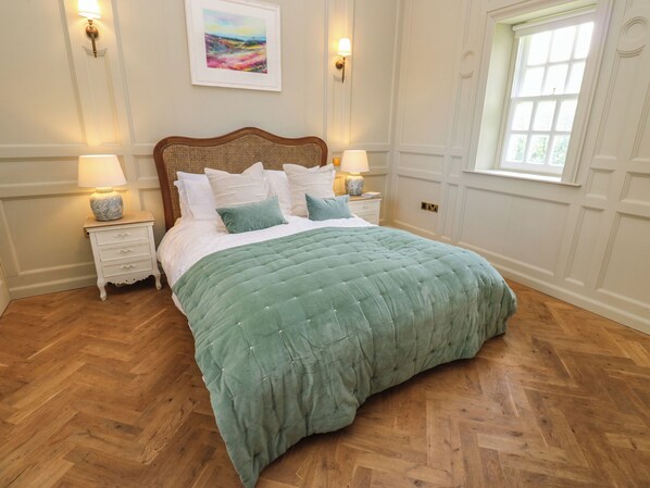 A neatly made bed with a green quilt, two bedside tables with lamps, and a window with a view of greenery.