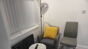 A room with a grey armchair, a small round table with a remote control, and a window with blinds. - Stunning Well Decorated 1bed Apartment in Dartford (Dartford)