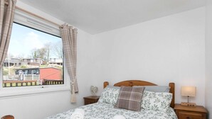 Un dormitorio con cama de madera, una ventana con cortinas, una mesita de noche con lámpara y vistas a una zona residencial.