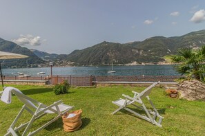 Un espacio verde con dos sillas de jardín blancas, una cesta tejida pequeña y una barandilla de metal con vistas a un lago con barcos y un pueblo lejano.