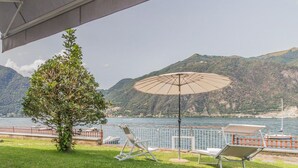 Uma área de estar externa com uma mesa posta para dois, com vista para um lago e montanhas ao fundo.