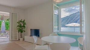 A modern living room with a large window offering a view of a lake and mountains. There is a round table with white chairs, a potted plant, and a flat-screen TV on a white cabinet. - Ninfea Apartments by Wonderful Italy - Arctic (Lezzeno)