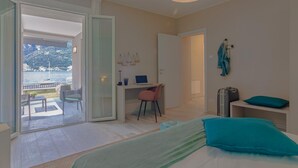 A bedroom with a bed, a desk, a chair, and a suitcase. There is a view of a balcony with outdoor furniture and a body of water in the background. - Ninfea Apartments by Wonderful Italy - Arctic (Lezzeno)