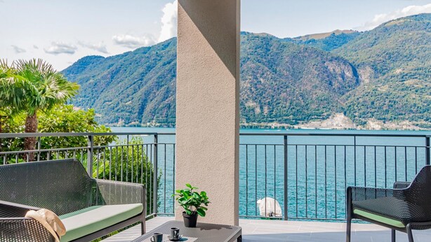 A balcony with two chairs, a small table, and a potted plant. The balcony overlooks a body of water and mountains.