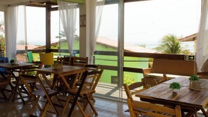 A restaurant with wooden tables and chairs, large windows with curtains, and a view of the ocean and buildings. - Pousada Brisa da Canoa (Aracati)