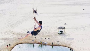 A person performing a high-flying trapeze act in an open area with a small body of water and a few people on the ground. - Pousada Brisa da Canoa (Aracati)