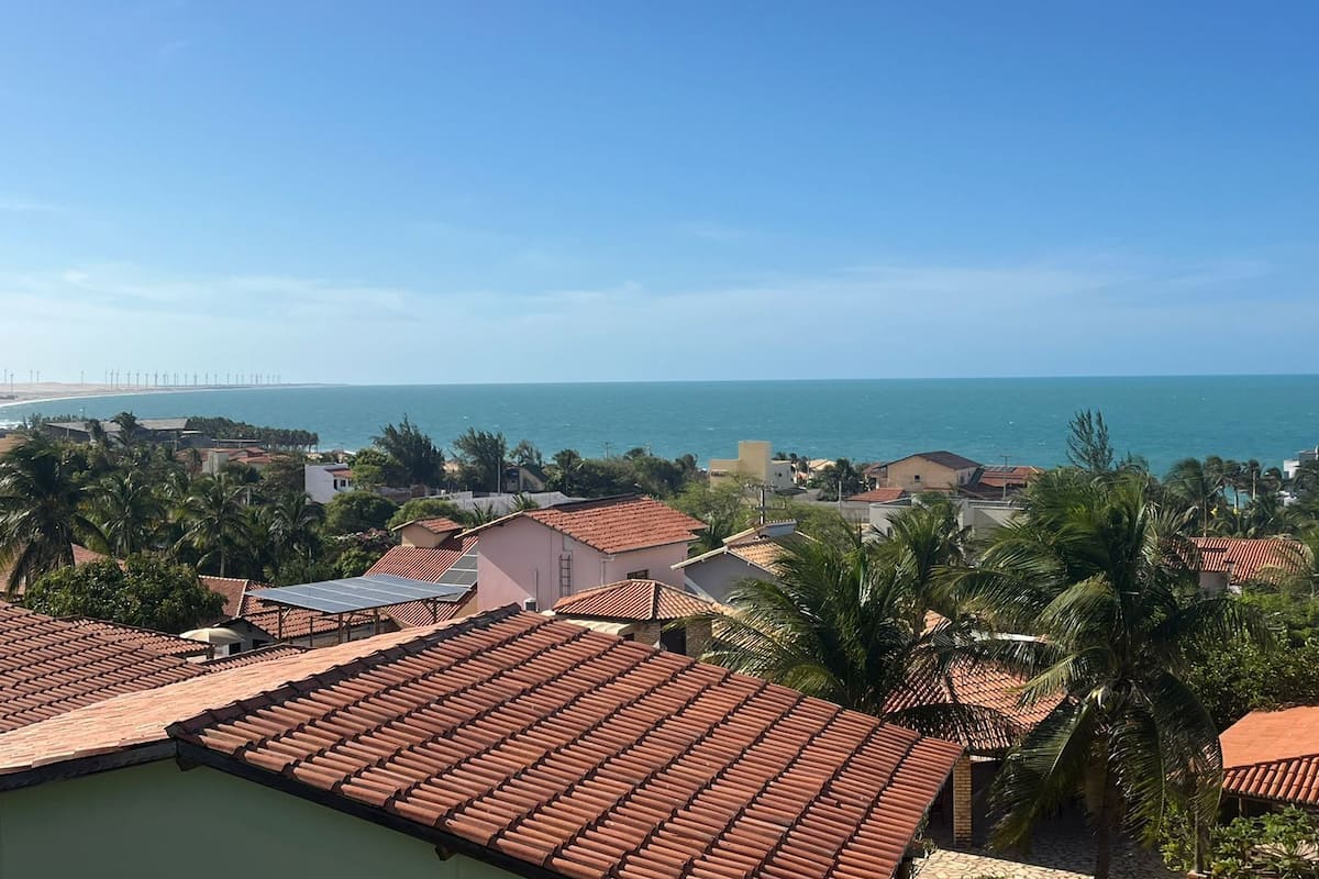 A coastal residential area with terracotta-tiled roofs, palm trees, and a view of the sea and a distant bridge.
