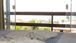 A bed with a white towel and a book, a balcony with a view of palm trees and buildings.