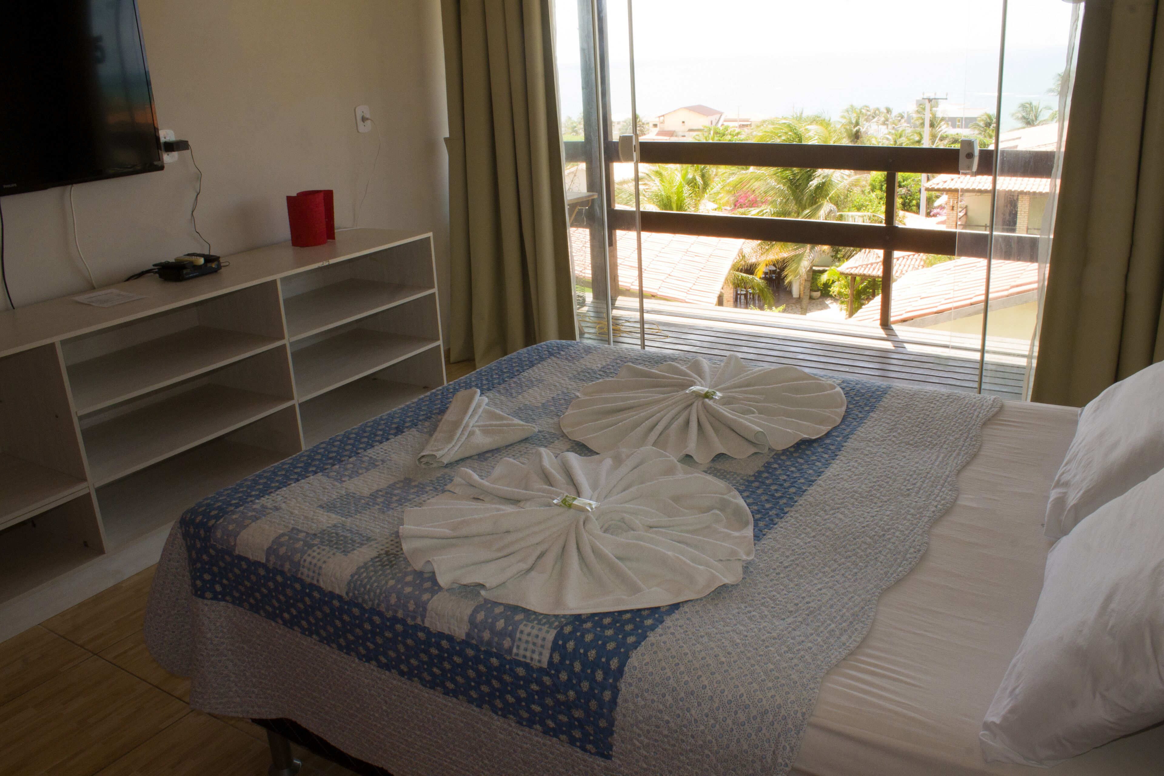 Een hotelkamer met een bed, een aan de muur bevestigde televisie, een raam met uitzicht op het omliggende landschap, en een rode beker op een plank.